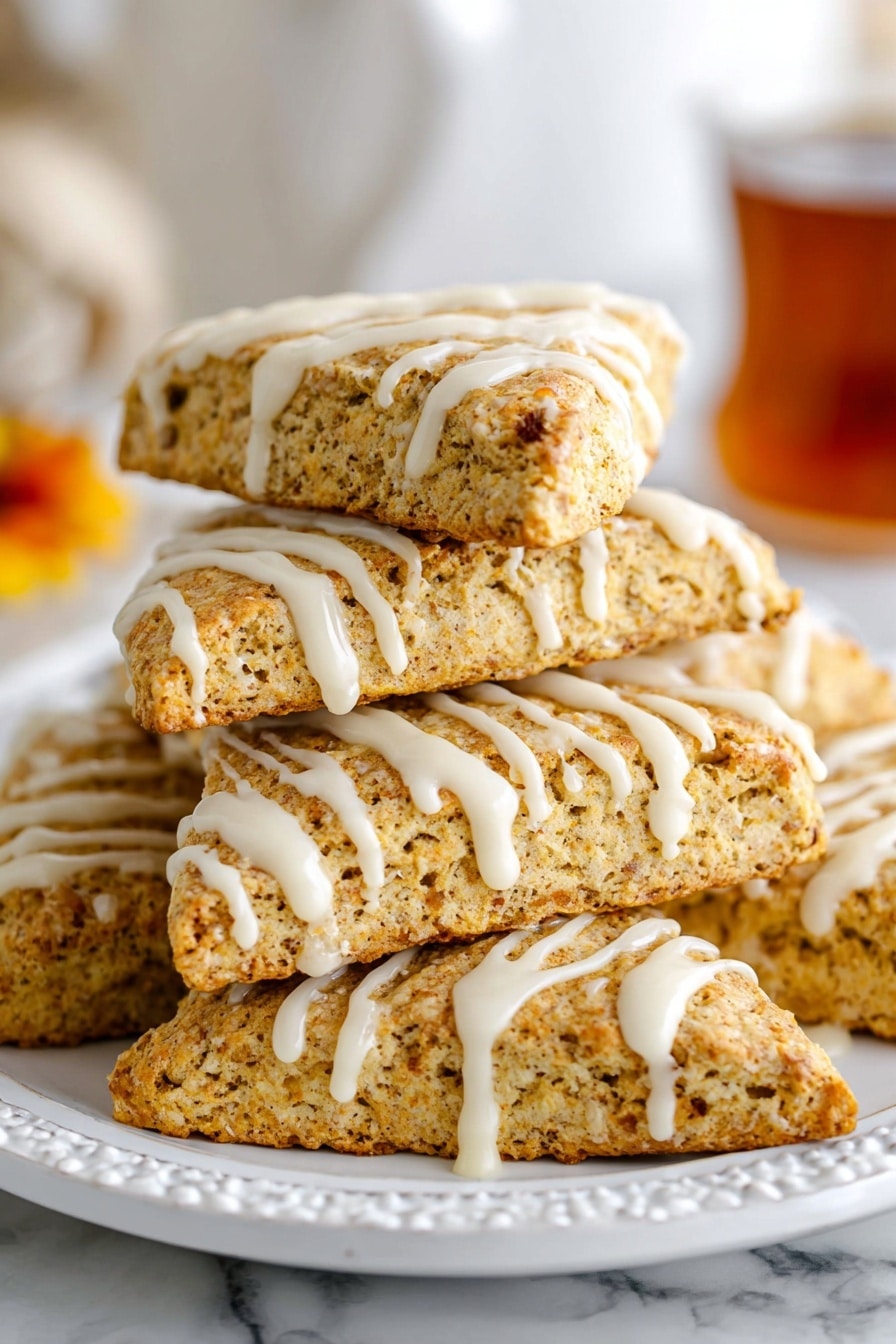 A triangular golden-brown scone sits on a white plate, with creamy white icing drizzled unevenly across the top, creating a slightly thick, dripping effect on the sides. In the background, more scones with similar icing are stacked, showing their crumbly texture. Next to the white plate, there is a glimpse of a glass bottle labeled